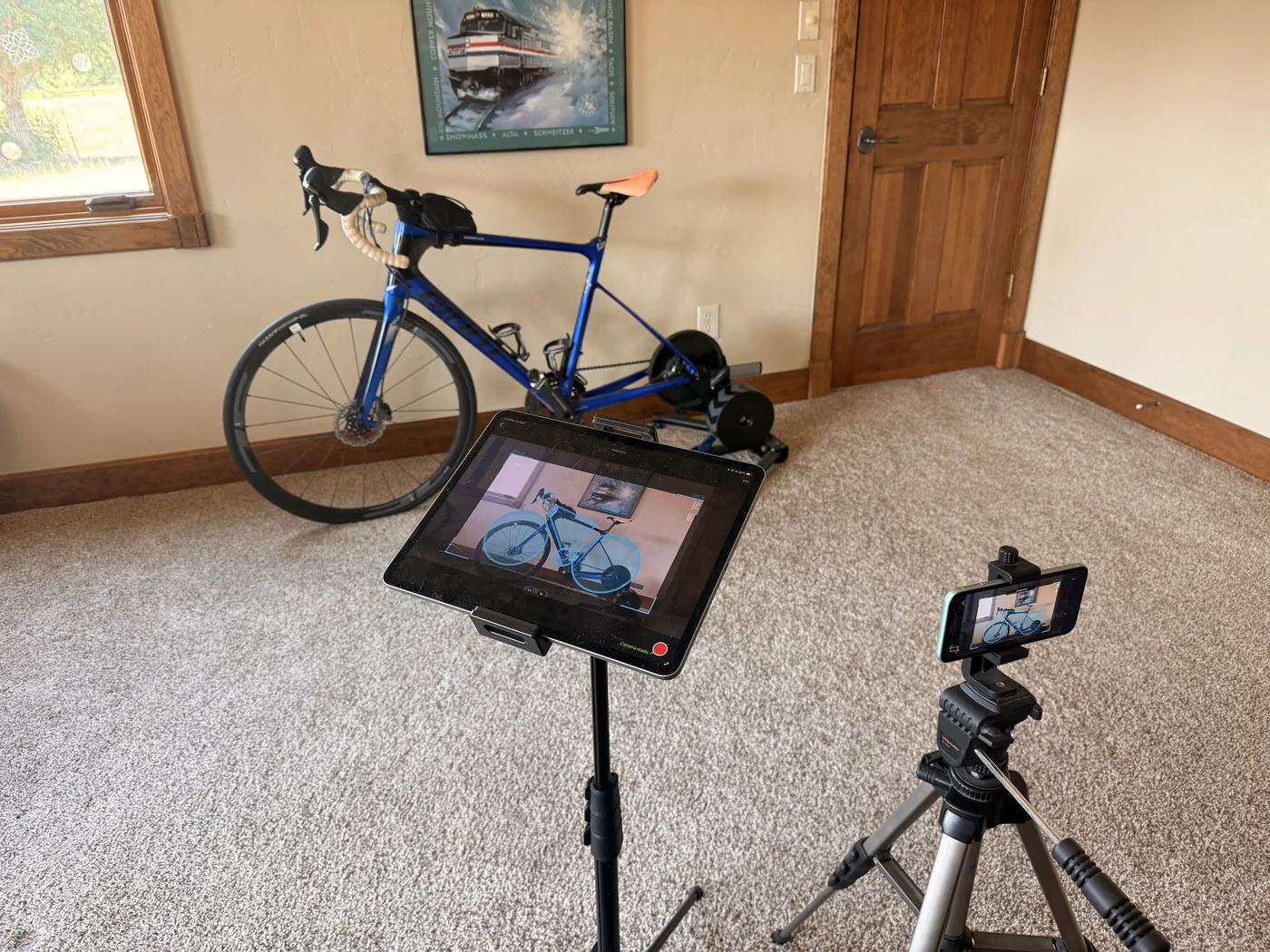 A road bike on an indoor trainer with an iPhone on a tripod filming it, and an iPad on a stand in front showing the live camera view of the bike